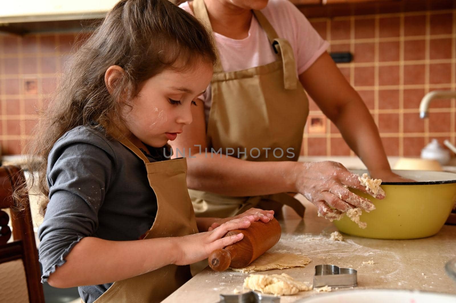 Side portrait of beautiful little girl, wearing a beige chef's apron, kneading and rolling dough with a wooden rolling pin. Cutting molds and fresh ingredients on a kitchen countertop. Baking concept