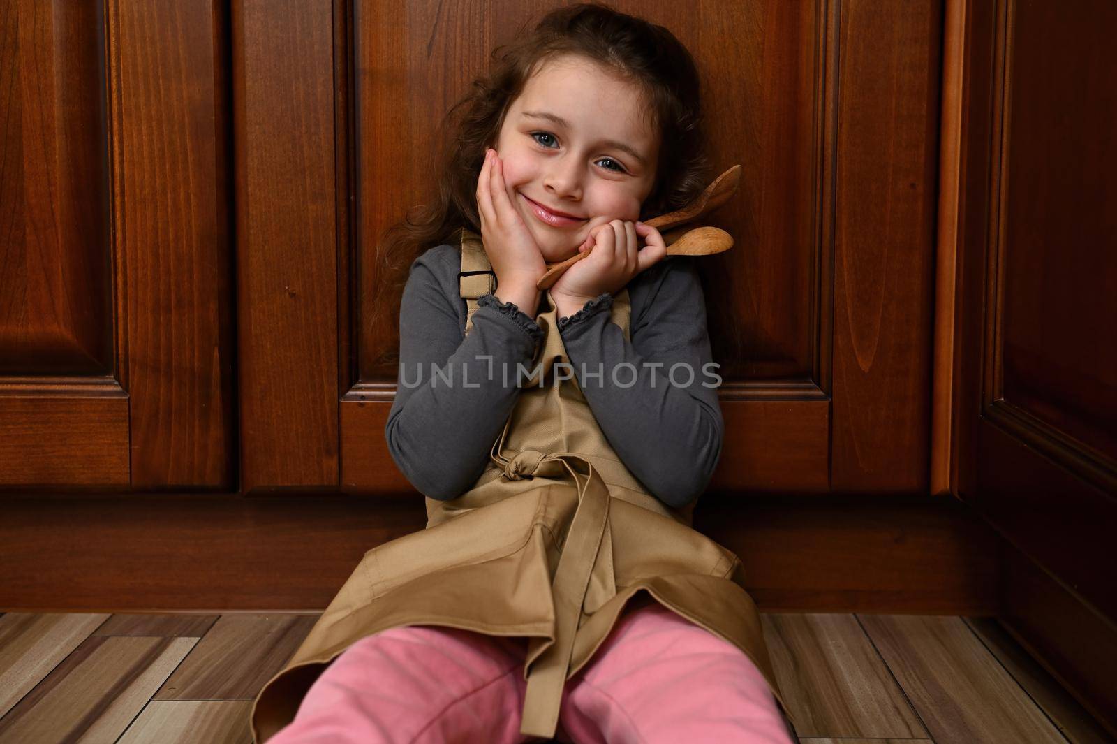 Adorable little girl in beige chef apron, sitting on the floor, leaning against wooden kitchen cupboard, holding cooking utensils, cutely smiling, looking to camera. Confectioner child, baker, cook