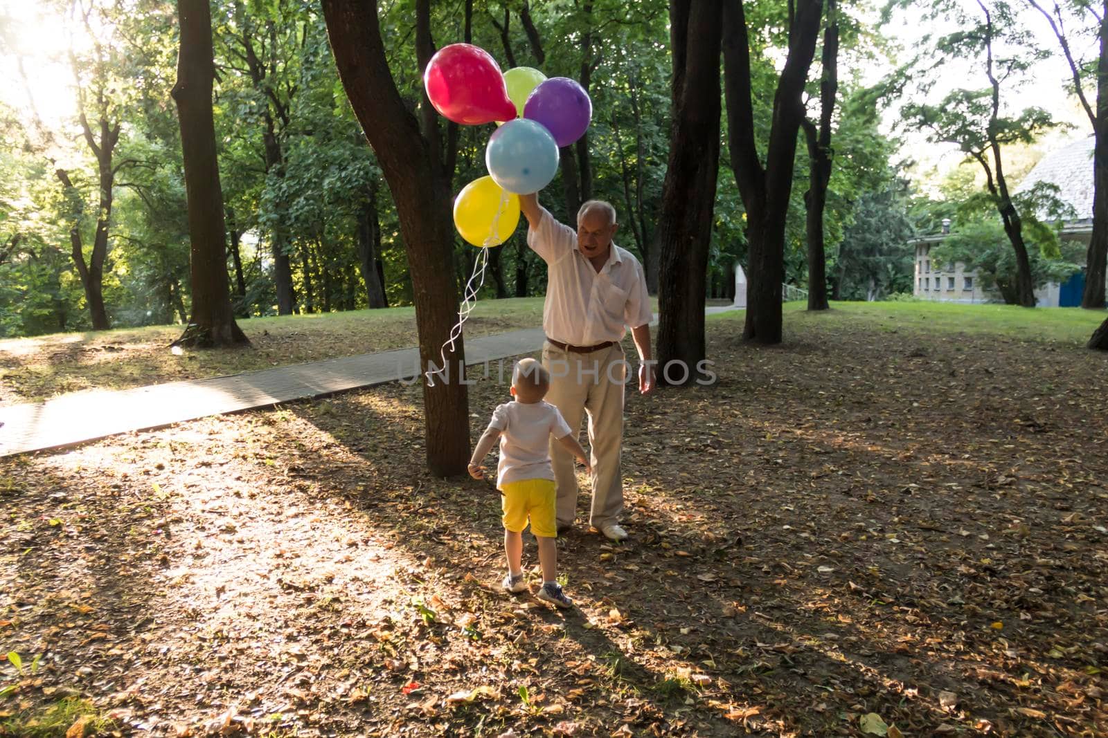 A little boy in yellow shorts and an elderly old man are playing with bright balloons. The family is having fun in the park in the fresh air and celebrating the holiday..