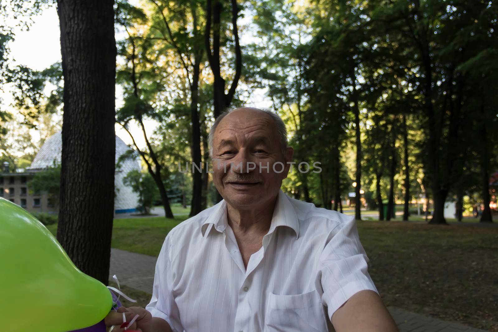 An elderly man with a smile on his face and bright balloons makes a selfie in the park.