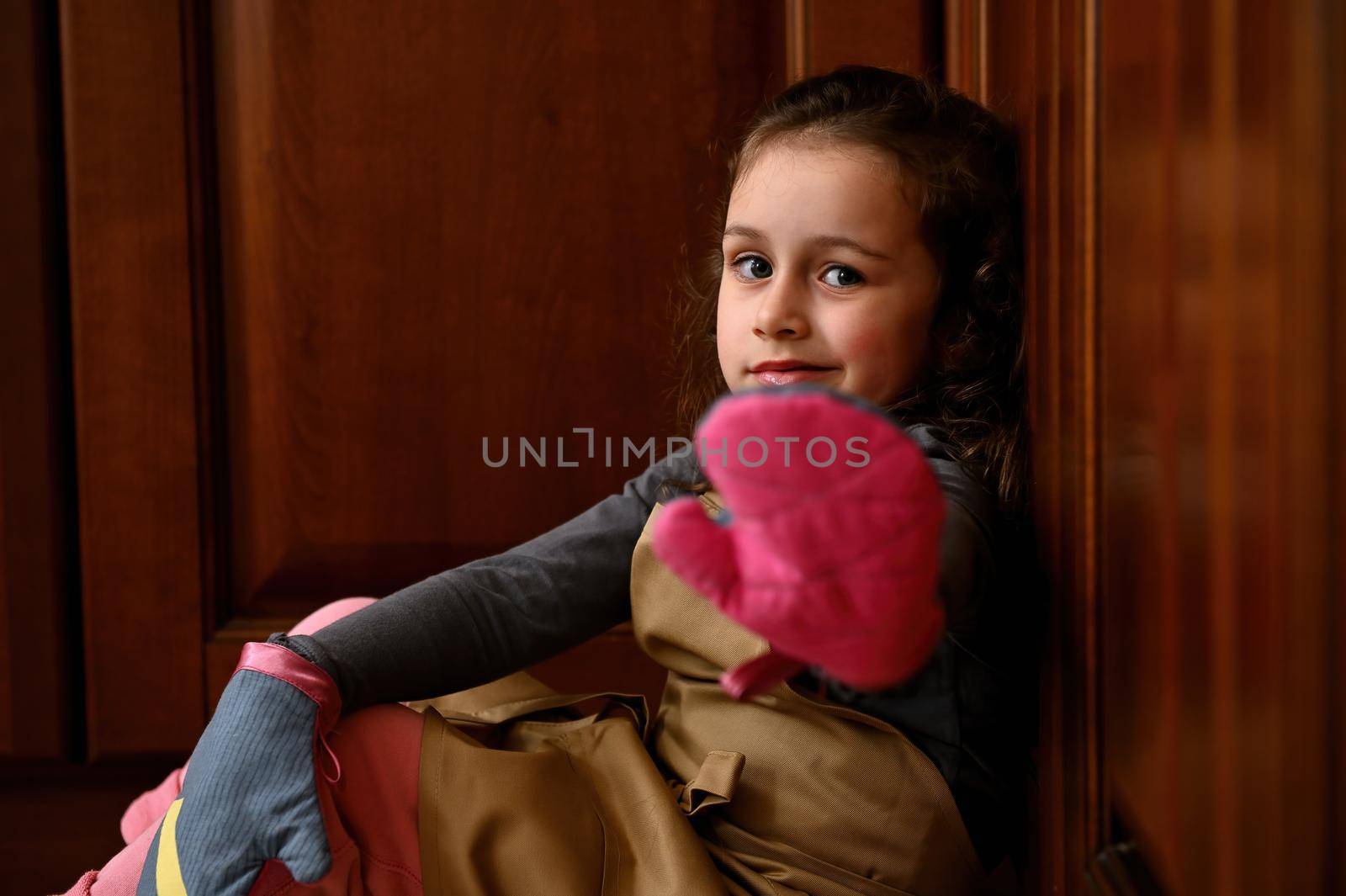 Caucasian little girl wearing beige chef's apron, sittting on the floor, leaning against wooden kitchen cupboard, handing her hand in mittens while looking to camera. Confectioner child, baker, cook