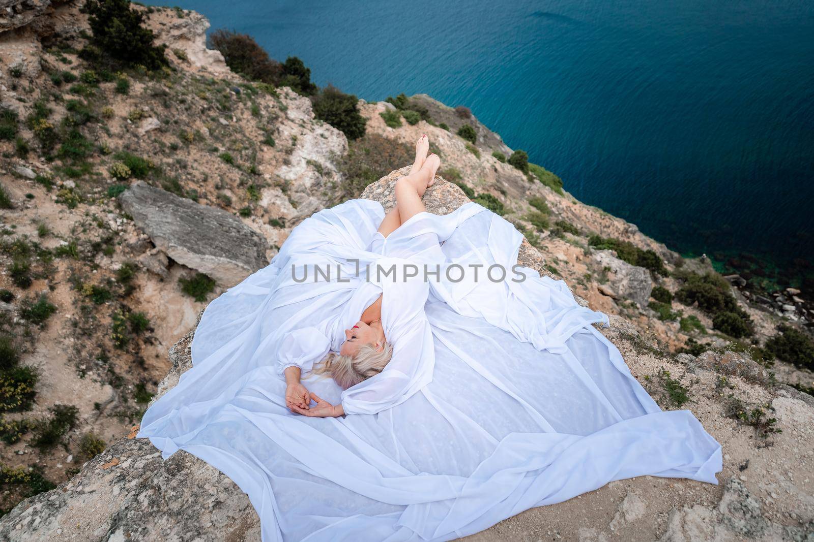 The blonde lies on a rock above the sea. With long hair on a sunny seashore in a white dress, rear view, silk fabric flutters in the wind. Against the backdrop of blue skies and mountains. by Matiunina