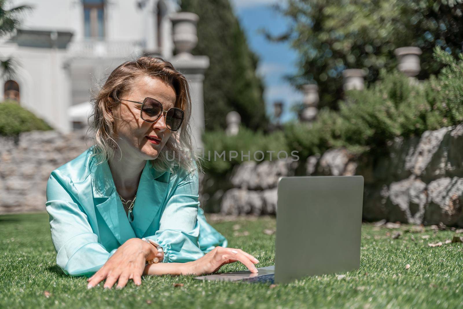 a young beautiful woman with blond curly hair in glasses and a blue dress sits on the grass in nature and uses a laptop