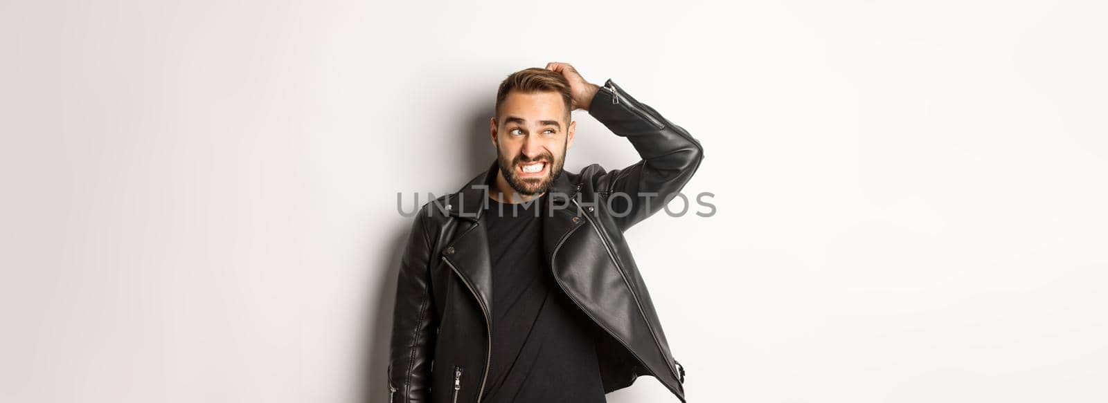 Confused macho guy in black leather jacket looking awkward and unsure, scratching head puzzled and stare left, white background.