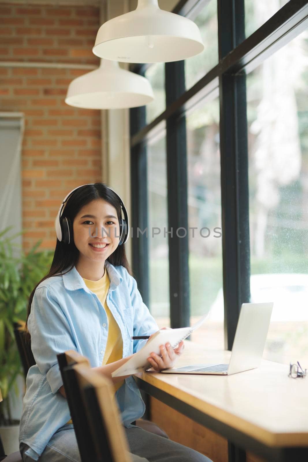 Happy young woman learning and studying online using laptop while sitting at table and looking at camera. Student learning online using laptop and smiling at the camera holding paperwork and sitting at table near window. Learning concept.
