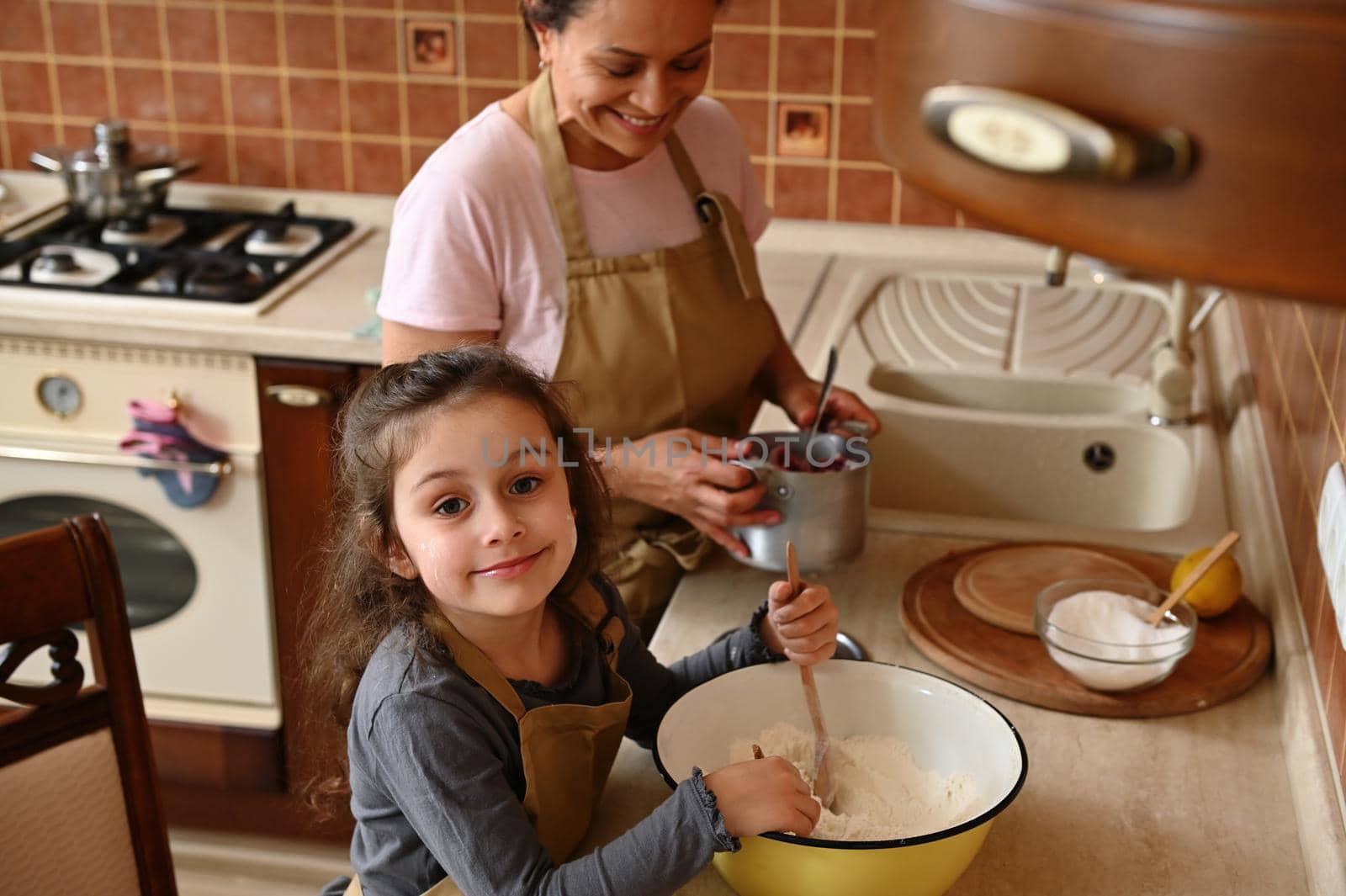 Beautiful child, European little girl in chef's apron, standing by a kitchen countertop near her mom and kneading dough. Happy childhood. Motherhood, Family pastime and relationships. Togetherness