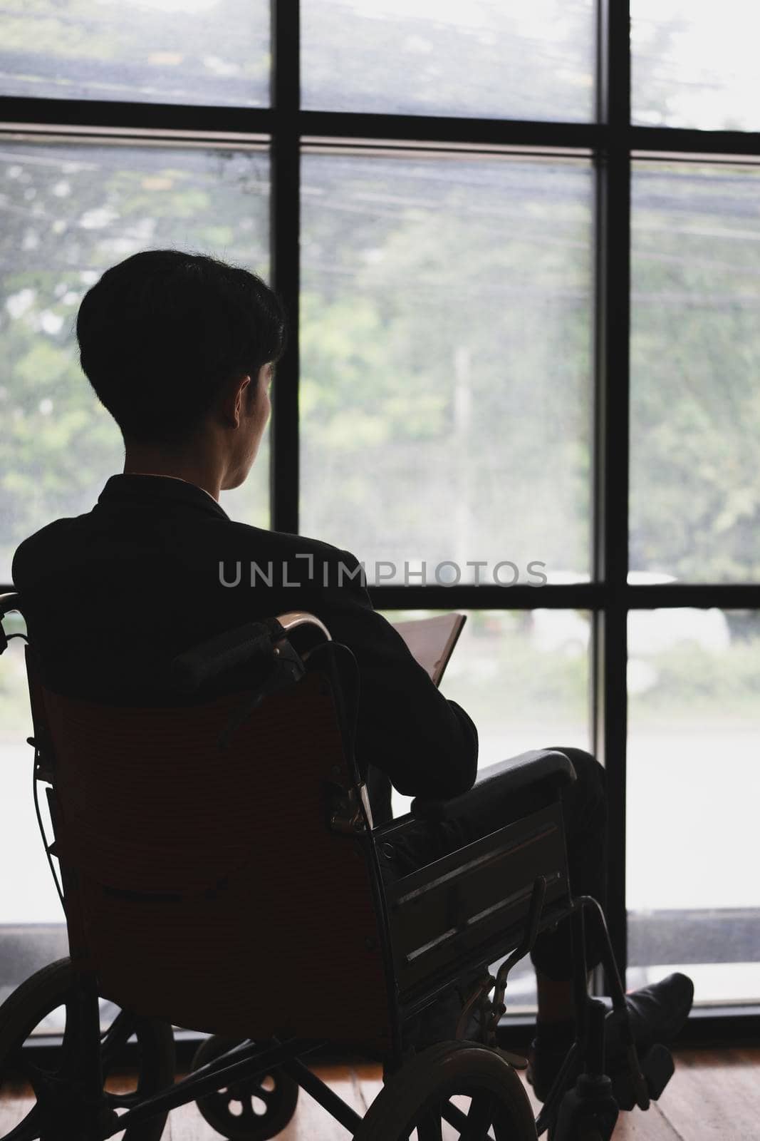 Rear view of thoughtful businessman in wheelchair sitting in office room and looking out of window. by prathanchorruangsak