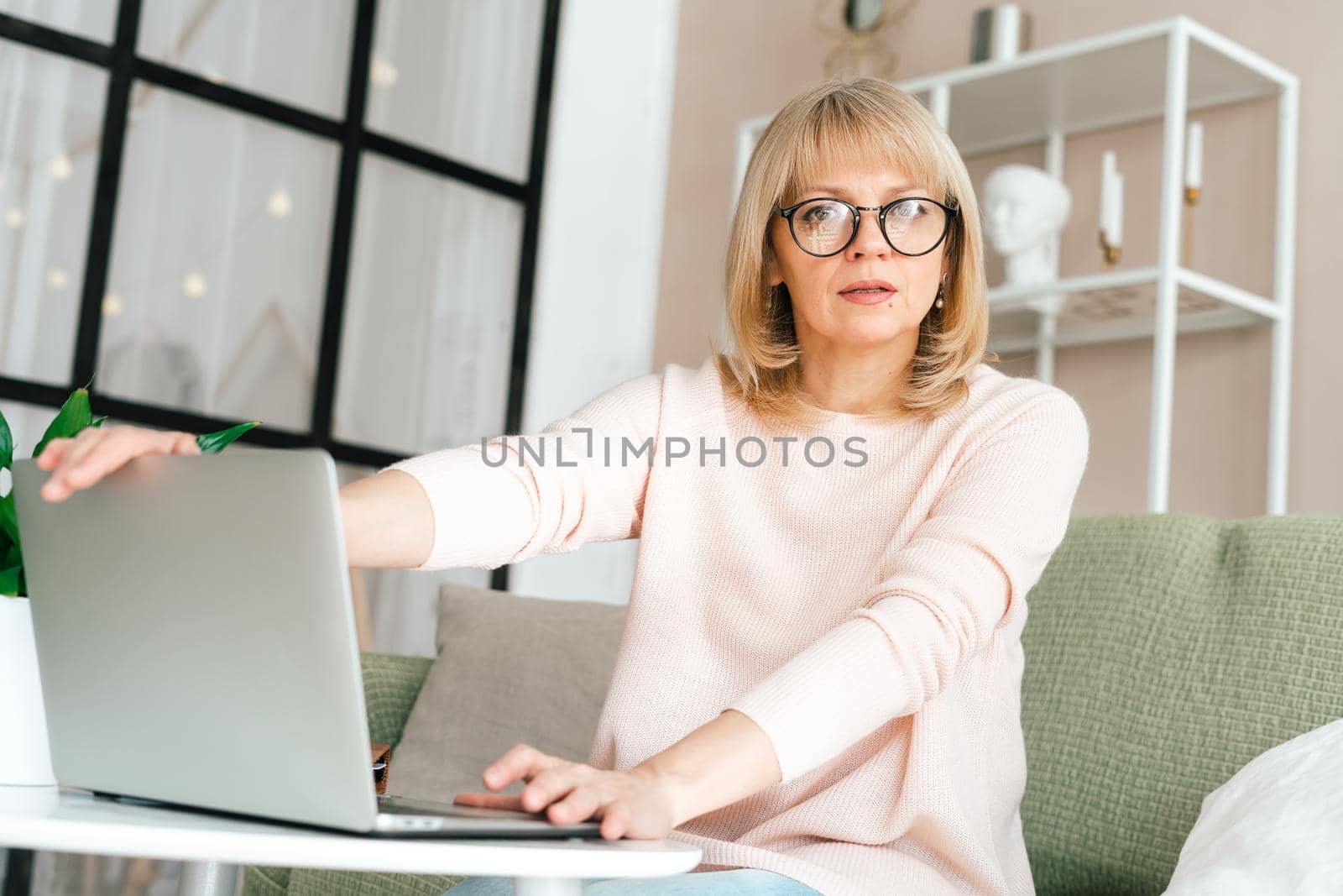 Thinking Freelance mature senior woman in glasses typing at laptop and working from home office with plants. Happy girl on workplace at the desk. Distance learning online education.