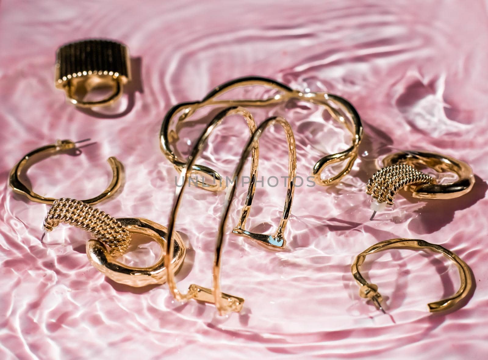 Golden bracelets, earrings, rings, jewelery on pink water background, luxury glamour and holiday beauty design for jewelry brand ads by Anneleven