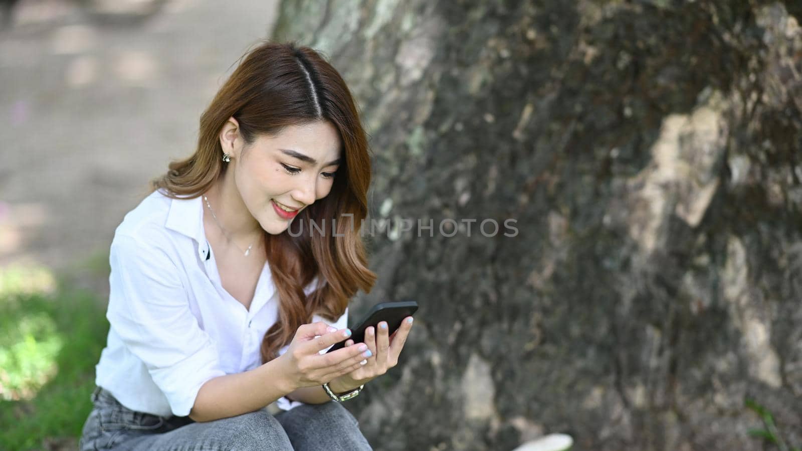 Beautiful young woman sitting under tree in the park and using smart phone. by prathanchorruangsak