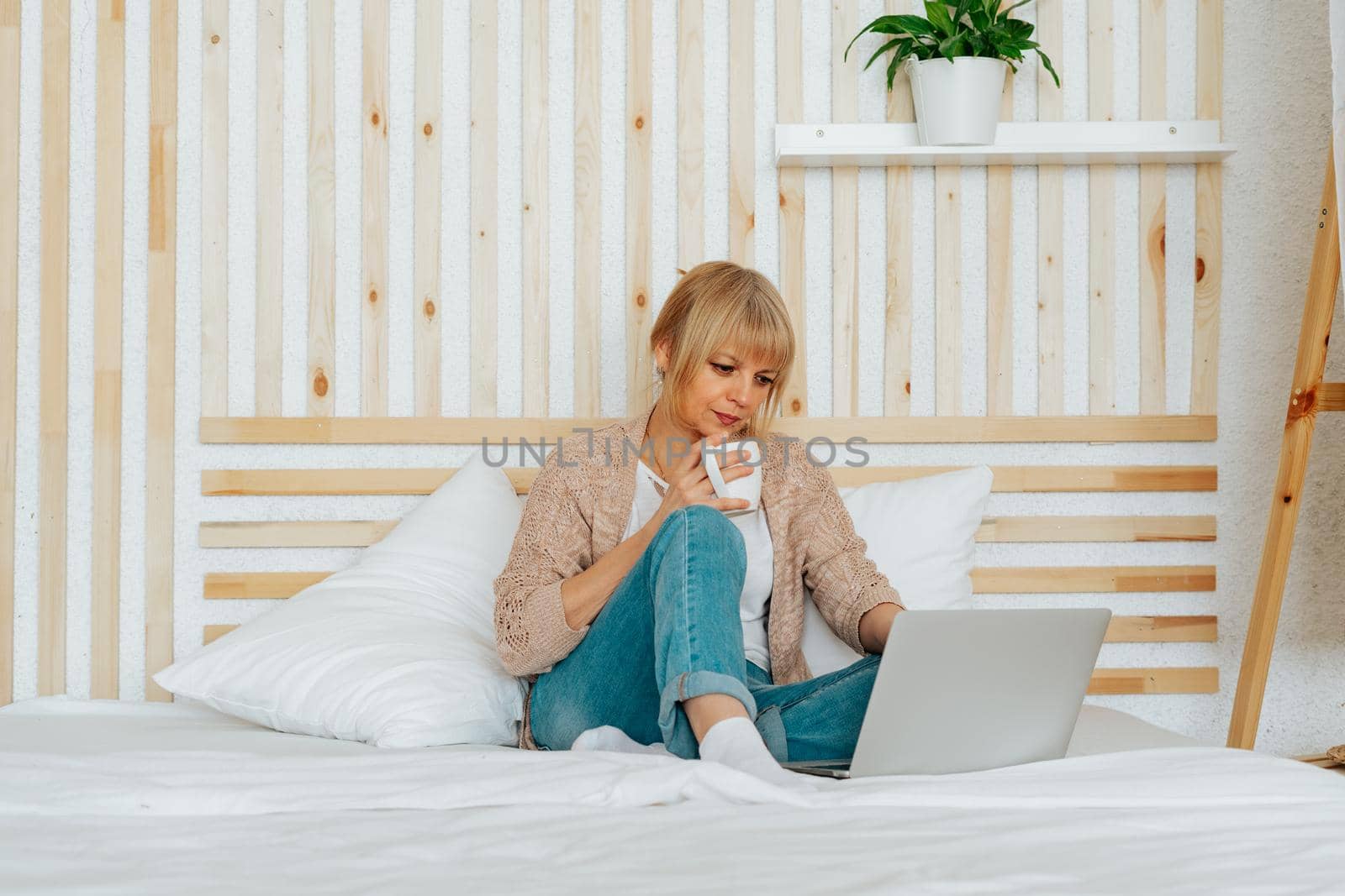 Senior mature woman sitting on bed with coffee mug and surfing browse internet on her laptop. Adult female lady using computer for online shopping banking video conference call while drinking tea.