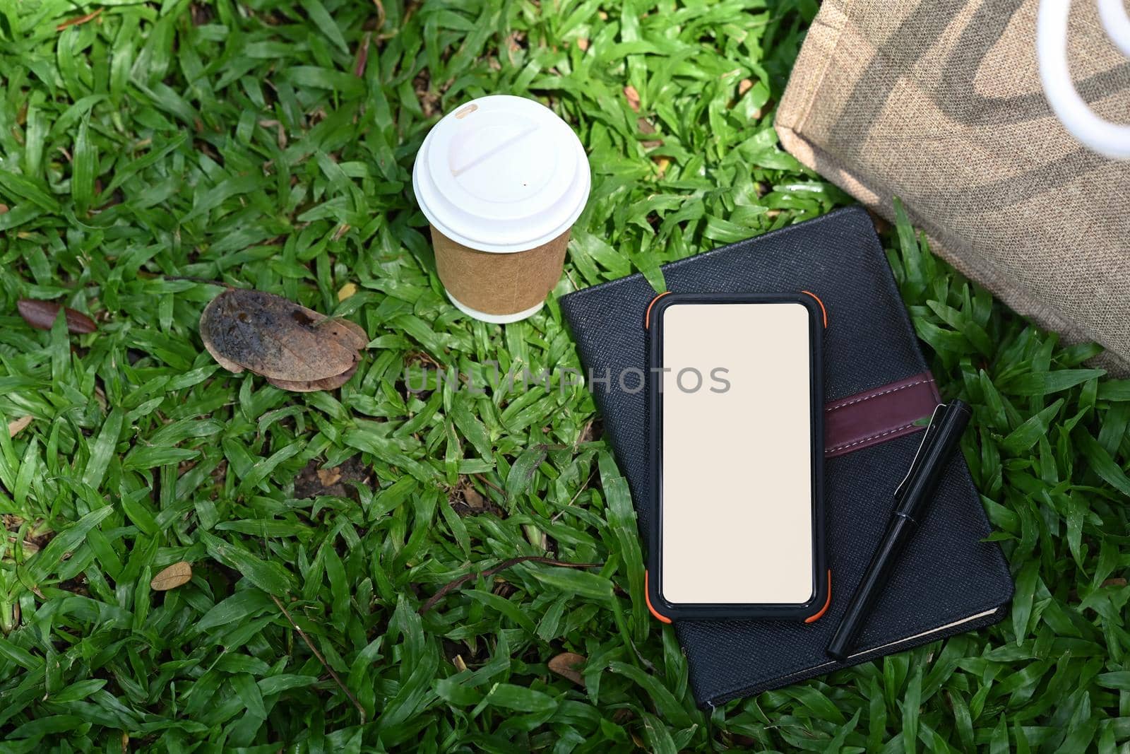 Mock up smart phone, coffee cup and book on green grass in the nature park.