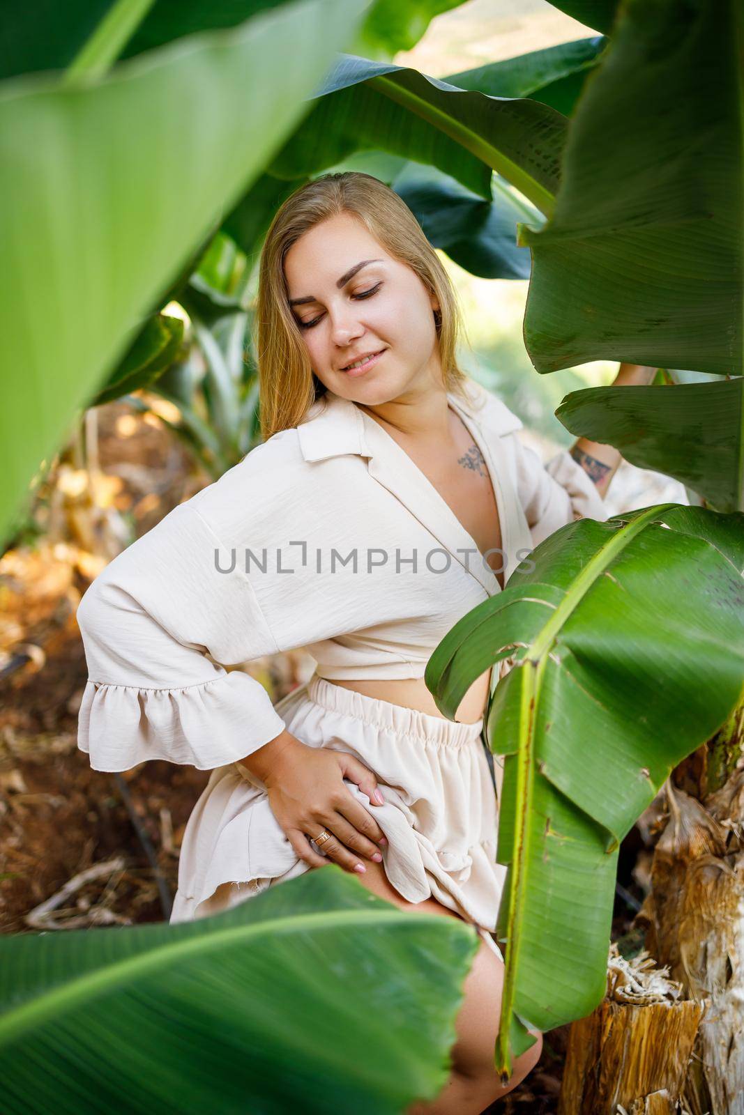 Woman near green leaves of banana bushes on nature in a park in a tropical place, she is in a beige skirt and a blouse by Dmitrytph