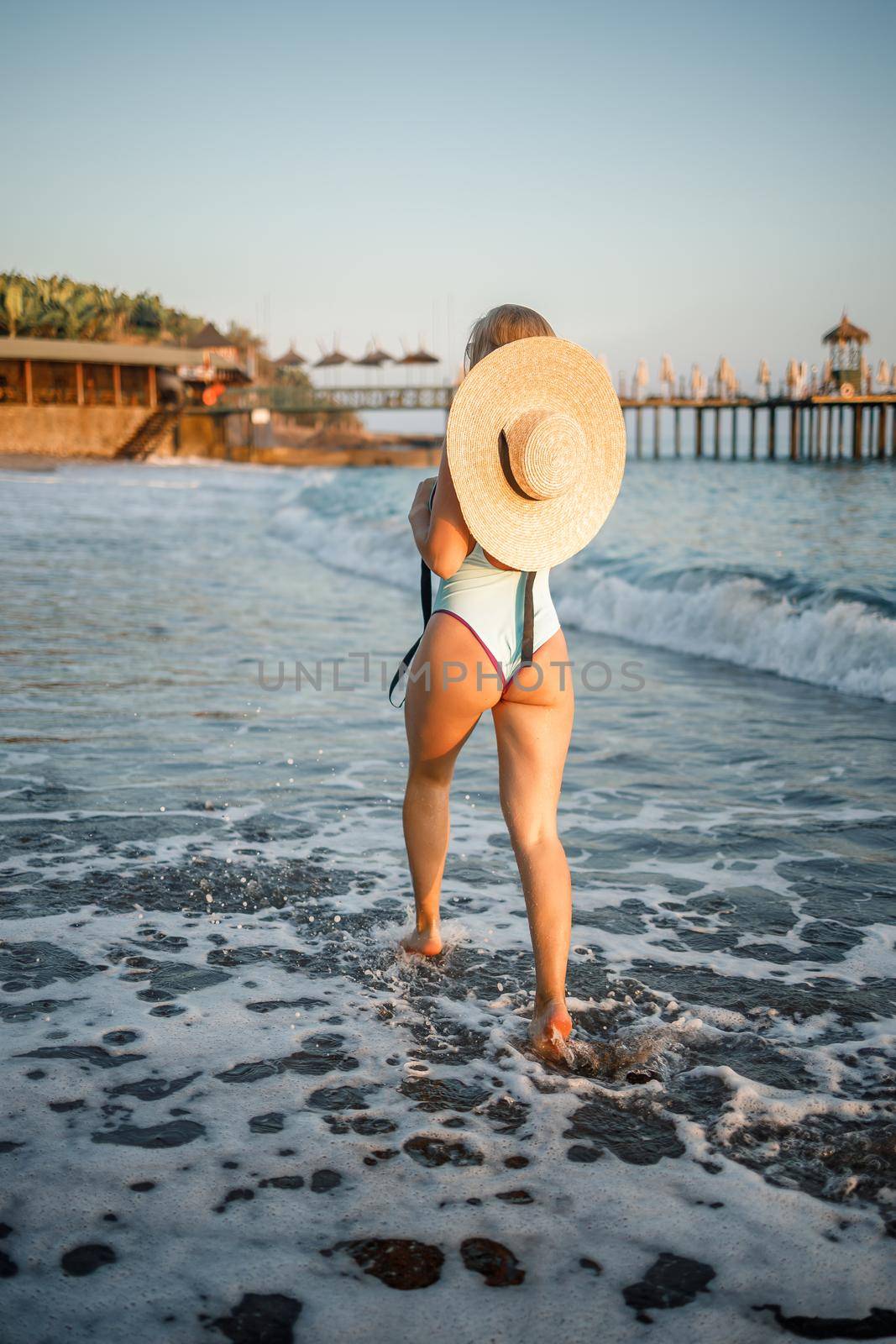 A woman in a swimsuit and a hat with glasses walks along the beach at sunset. The concept of sea recreation. Selective focus