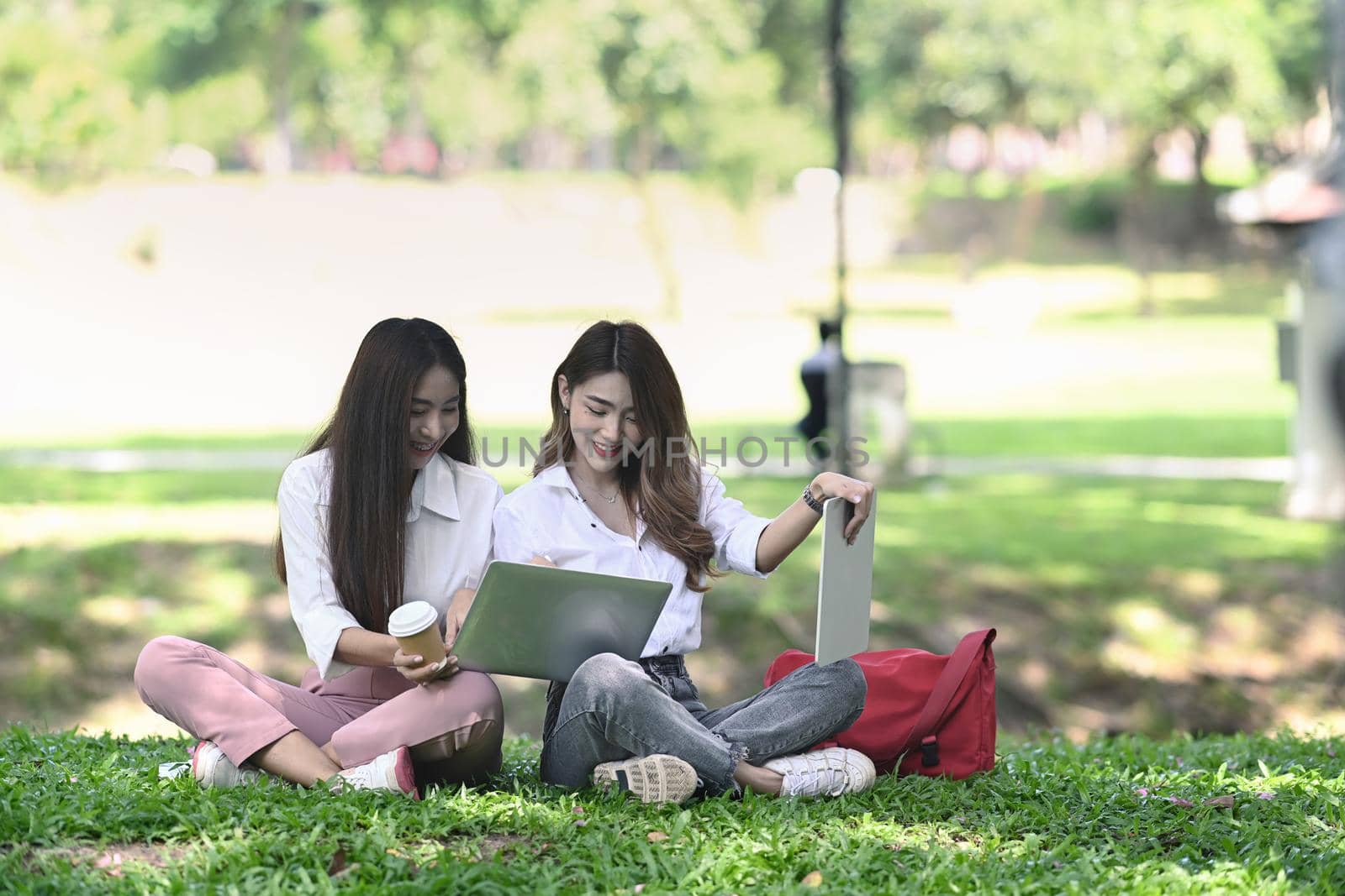 Two happy women sitting in the park and working together. by prathanchorruangsak