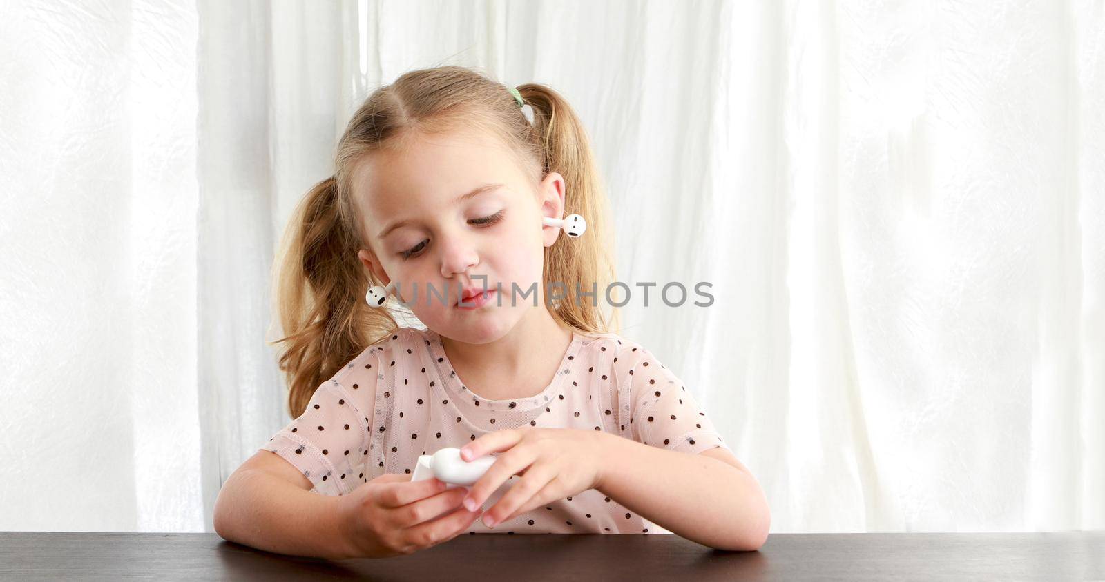 Cute little girl with true wireless earbuds inserted into ears in wrong manner playing with charging case at table