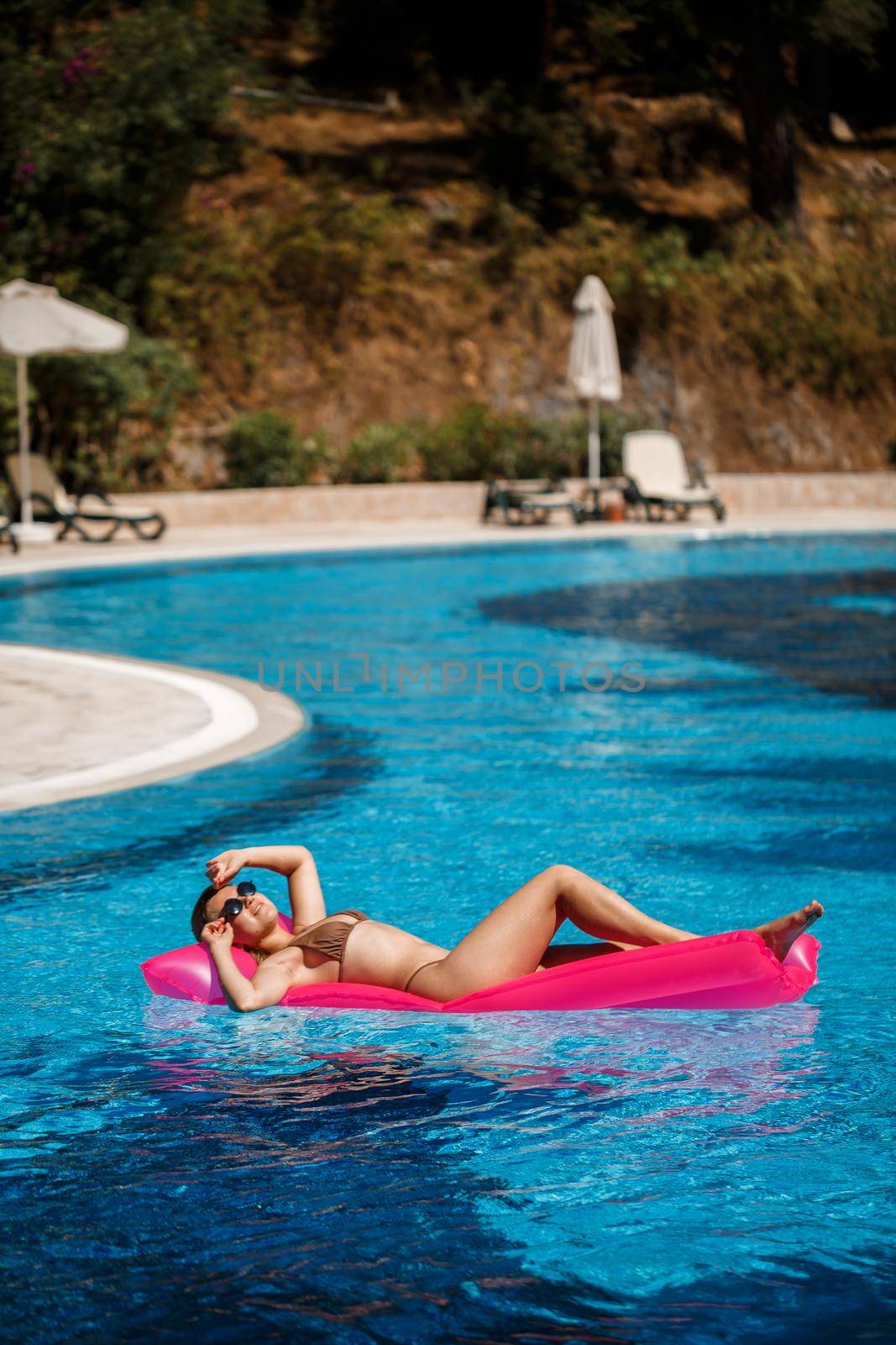 Sexy woman in a swimsuit lies on a pink inflatable mattress in the pool. Relax by the pool on a hot summer sunny day. Vacation concept