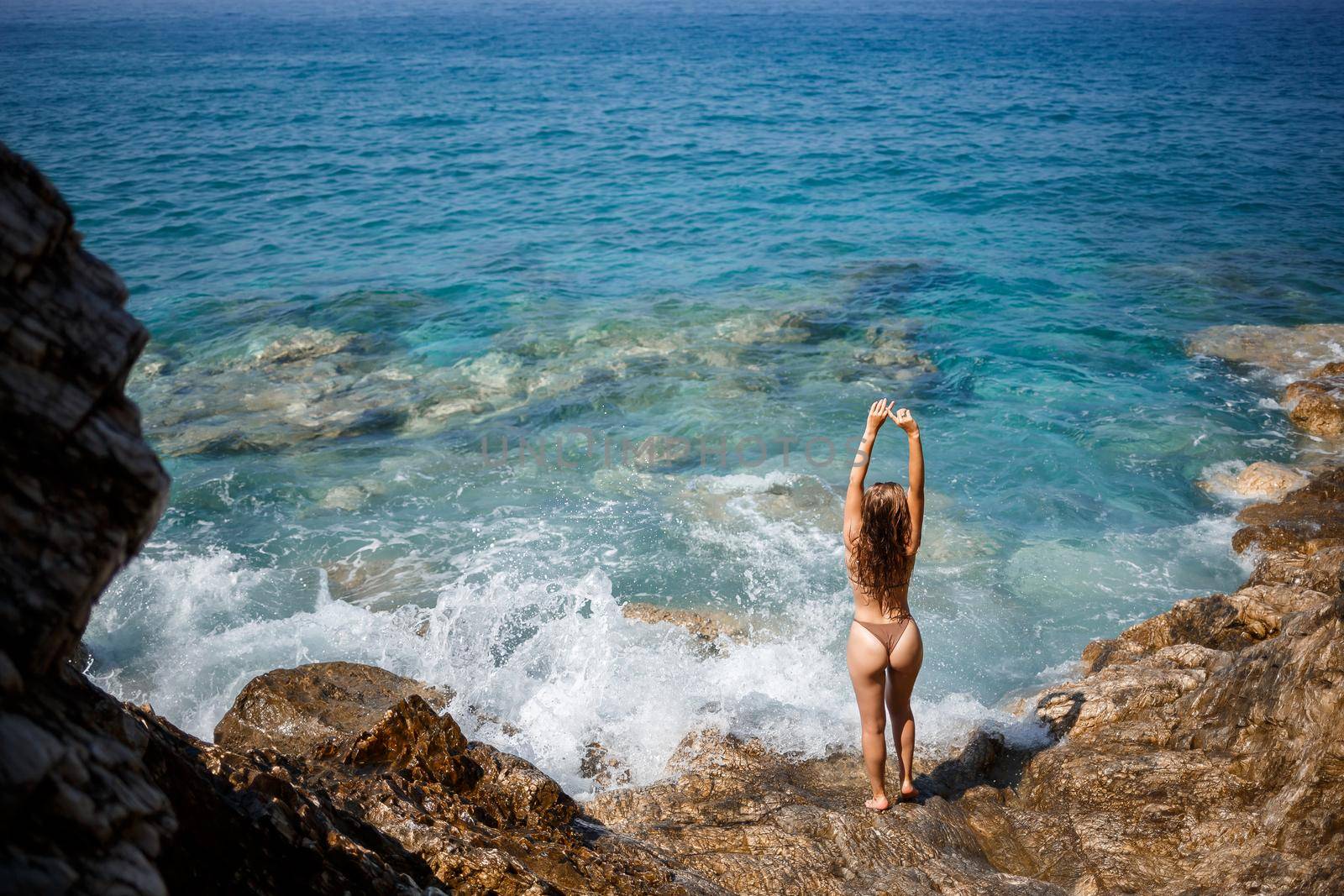 Sexy woman in full body swimsuit with long hair walks on large rocks on a rocky beach during a storm at sea. Back view. Woman swimwear island tropics landscape exotic walk