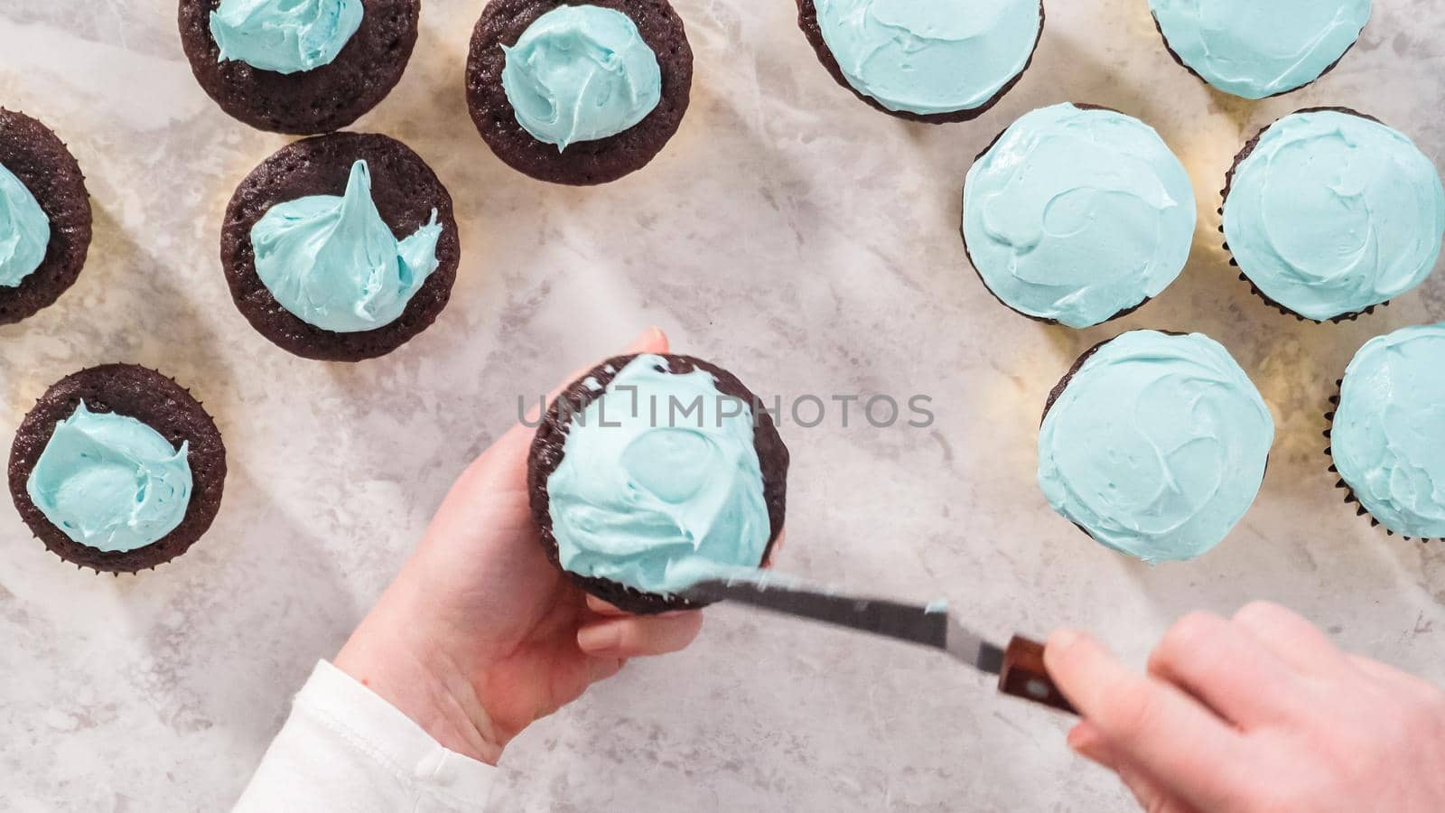 Flat lay. Step by step. Scooping blue buttercream frosting on top of the chocolate cupcakes.