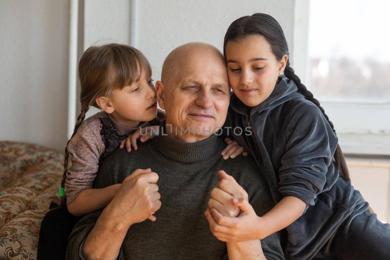 grandfather and two granddaughters hugging on sofa at home by Andelov13