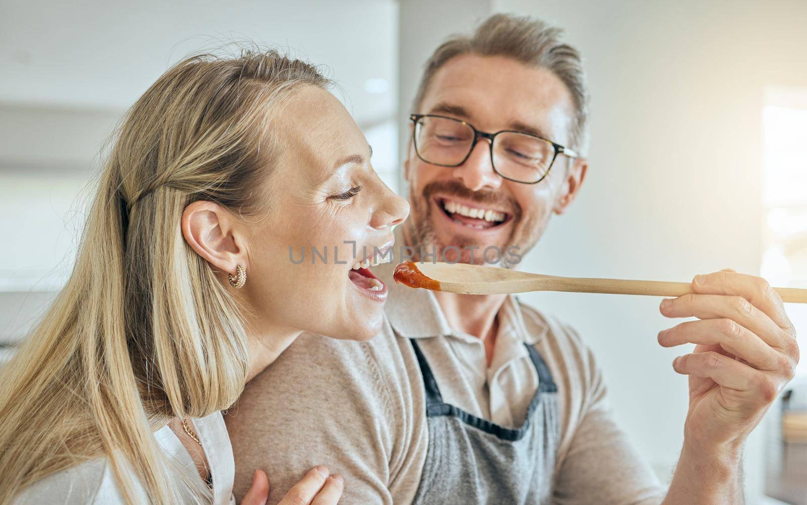 Happy couple cooking, tasting spoon of food and meal, lunch and dinner in kitchen at home. Smile mature man giving hungry woman a taste of homemade tomato sauce meal in mouth to enjoy eating together by YuriArcurs