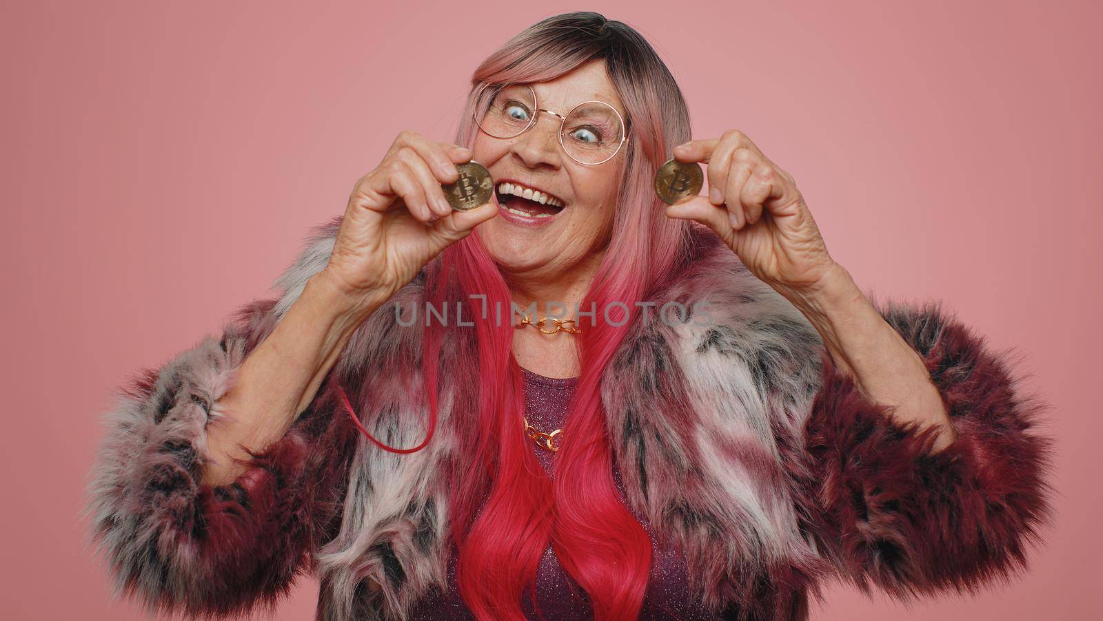 Senior elderly woman winner showing golden bitcoins BTC. Career wealth, cryptocurrency investment, mining future technology. Grandmother fooling making faces. Happy businesswoman on pink background