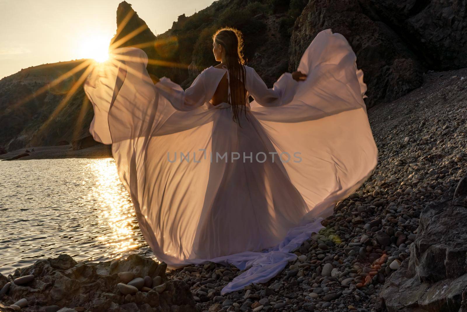 A mysterious female silhouette with long braids stands on the sea beach with mountain views, Sunset rays shine on a woman. Throws up a long white dress, a divine sunset. by Matiunina