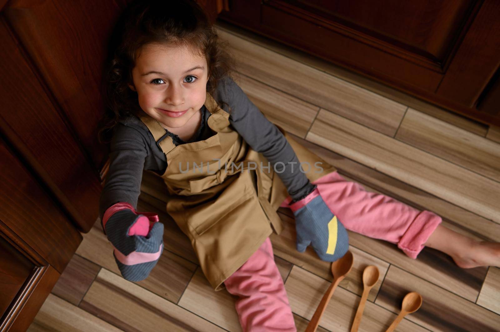 Little girl baker confectioner, chef pastry in chef apron and mittens shows thumbs up, sitting barefoot on kitchen floor by artgf