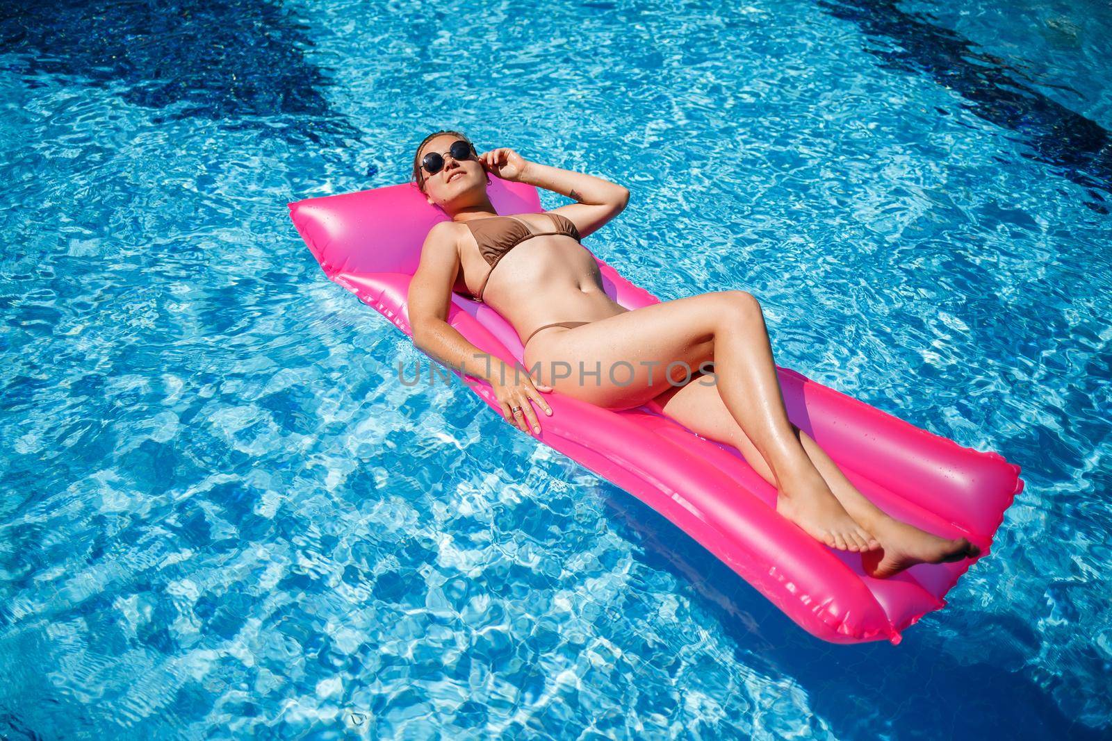 Sexy woman in a swimsuit lies on a pink inflatable mattress in the pool. Relax by the pool on a hot summer sunny day. Vacation concept