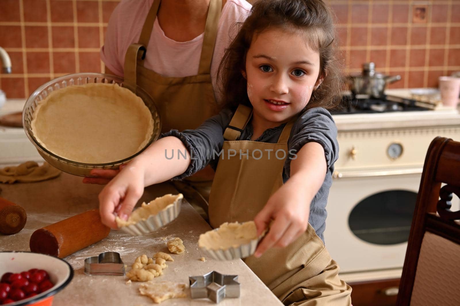 Mom and daughter in aprons, cooking together, showing rolled dough in the molds while making homemade tartlets and tarte by artgf