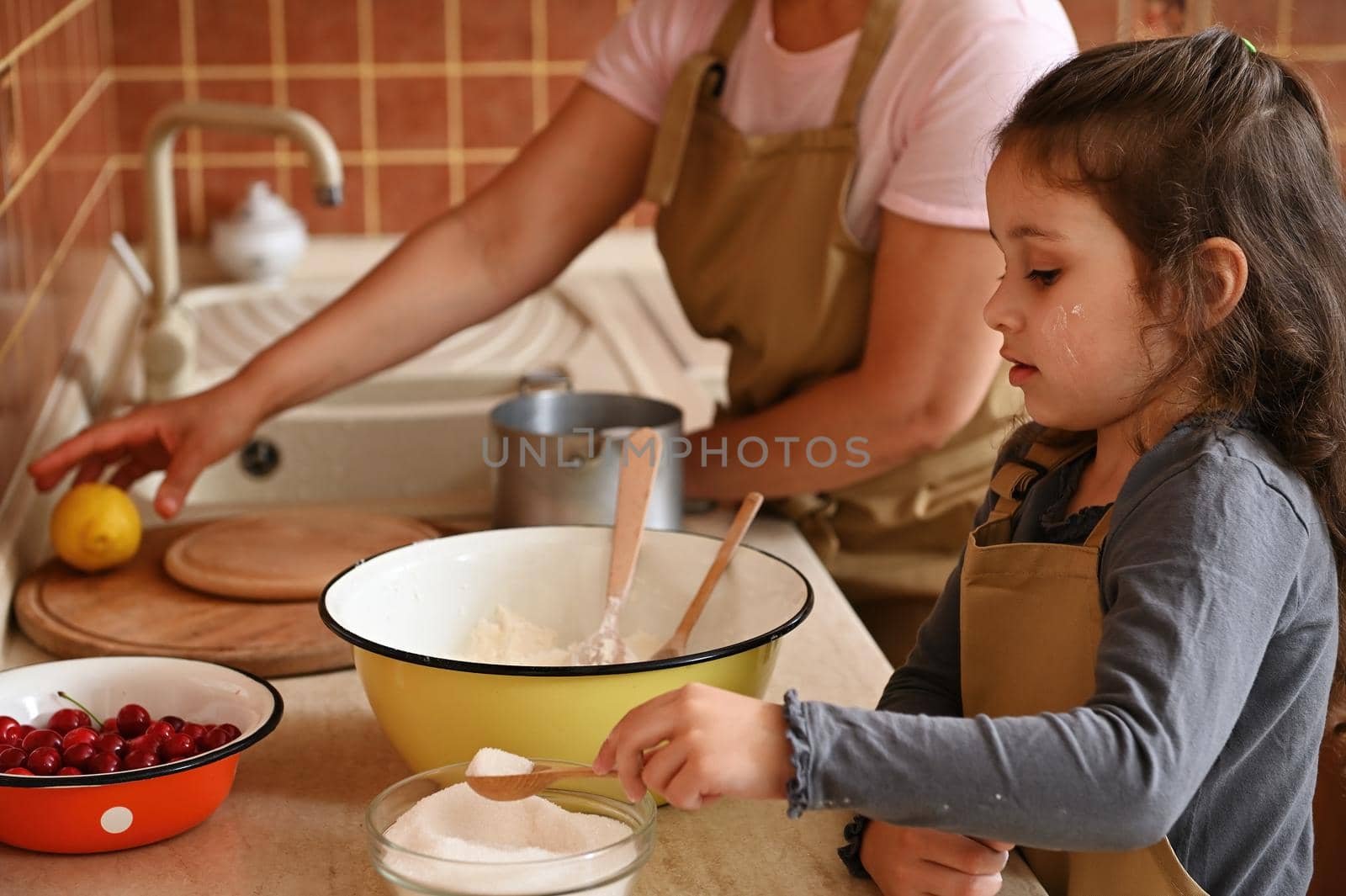 Beautiful little girl, adorable lovely daughter in chef's apron, adding scoop of sugar into a bowl with dry ingredients, preparing dough with her mother in home kitchen. Culinary and baking concept
