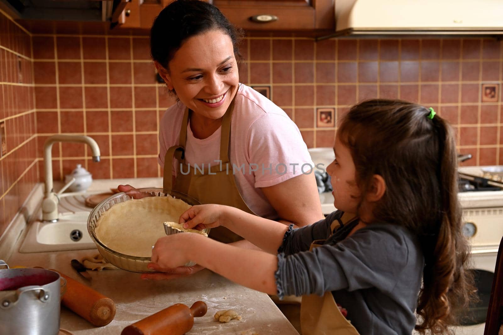 Mom and daughter smiling, laughing, playing, enjoying cooking together in kitchen, holding molds with rolled out dough by artgf