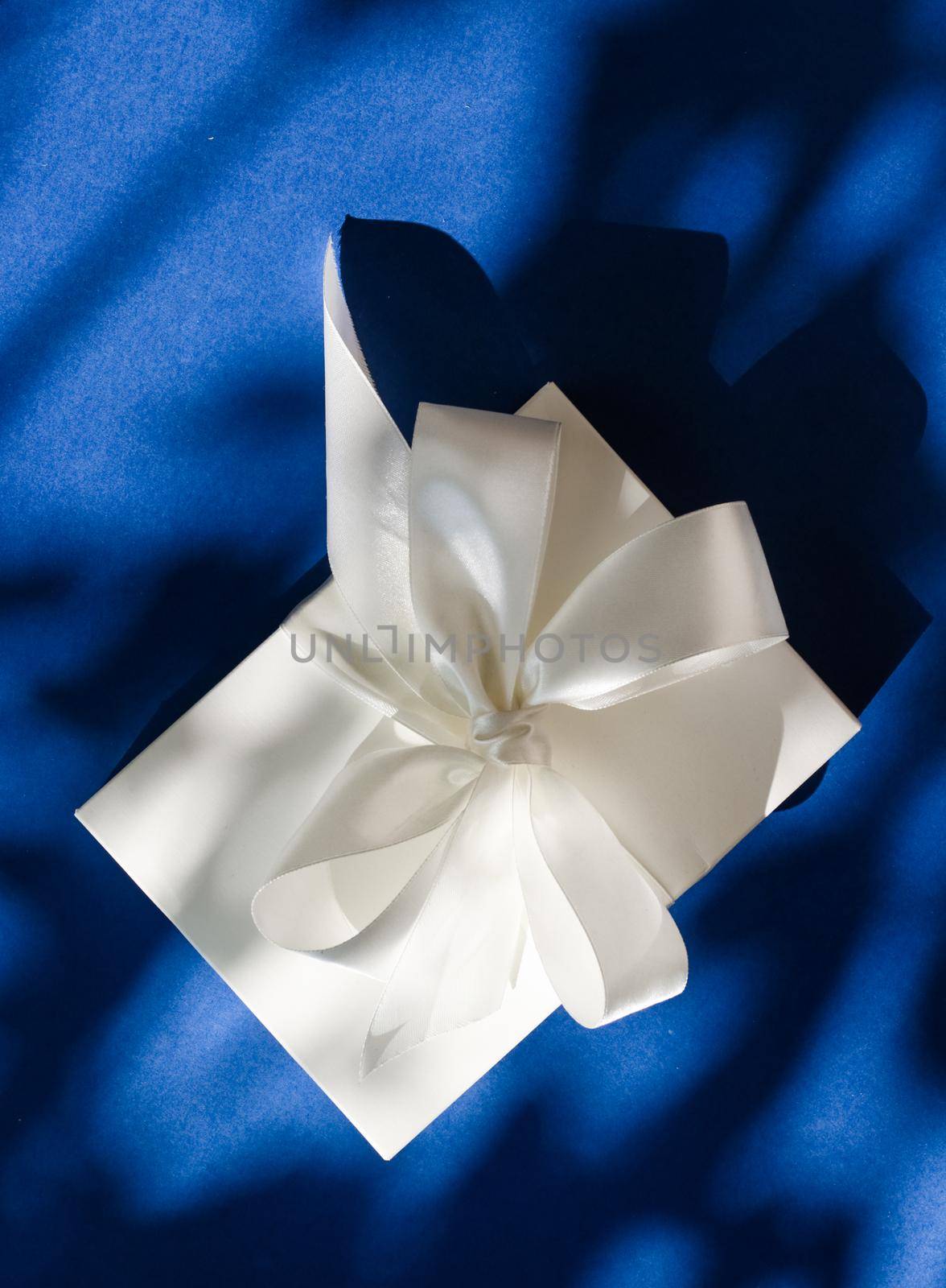 Luxury holiday white gift box with silk ribbon and bow on blue background, luxe wedding or birthday present by Anneleven