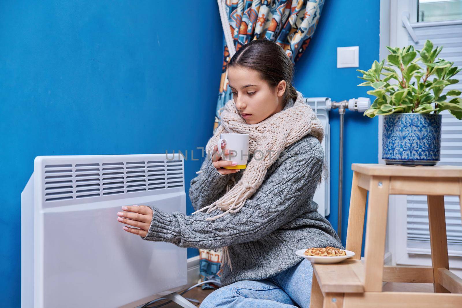 Cold autumn winter season, lifestyle, teenage female in warm sweater scarf warming near electric heating radiator, drinking hot drink with cup. Heating, warmth, equipment, life in cold season