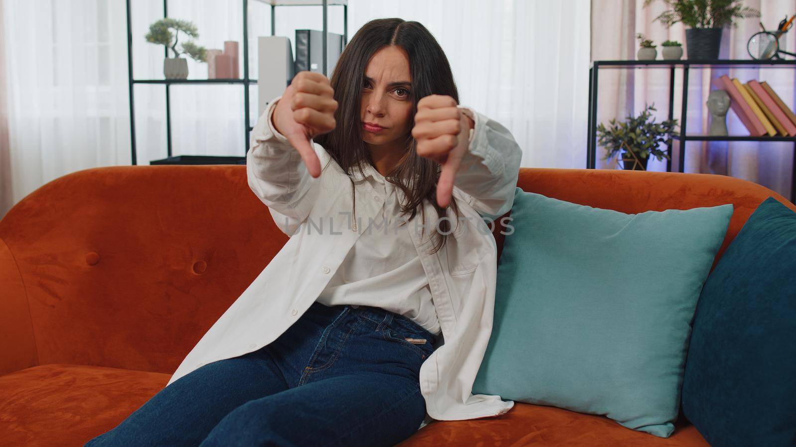 Dislike. Upset girl showing thumbs down sign gesture, expressing discontent, disapproval, dissatisfied bad work at modern home apartment indoors. Displeased young woman in living room sitting on sofa