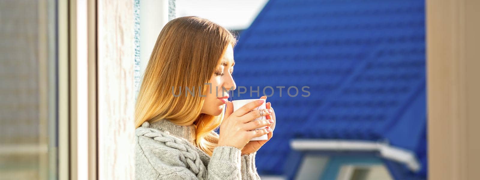 Woman relaxing on the balcony with coffee. A beautiful young woman in a sweater holds a cup of coffee standing near the window in the morning