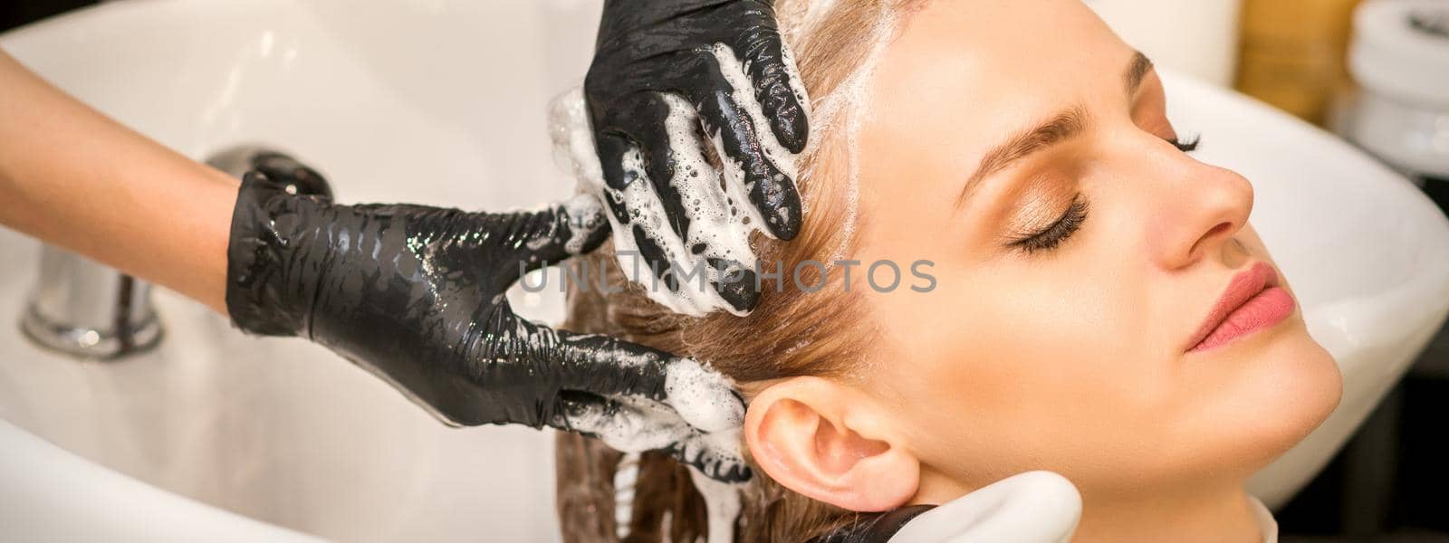 Young caucasian blonde woman having hair washed in the sink at a beauty salon. by okskukuruza