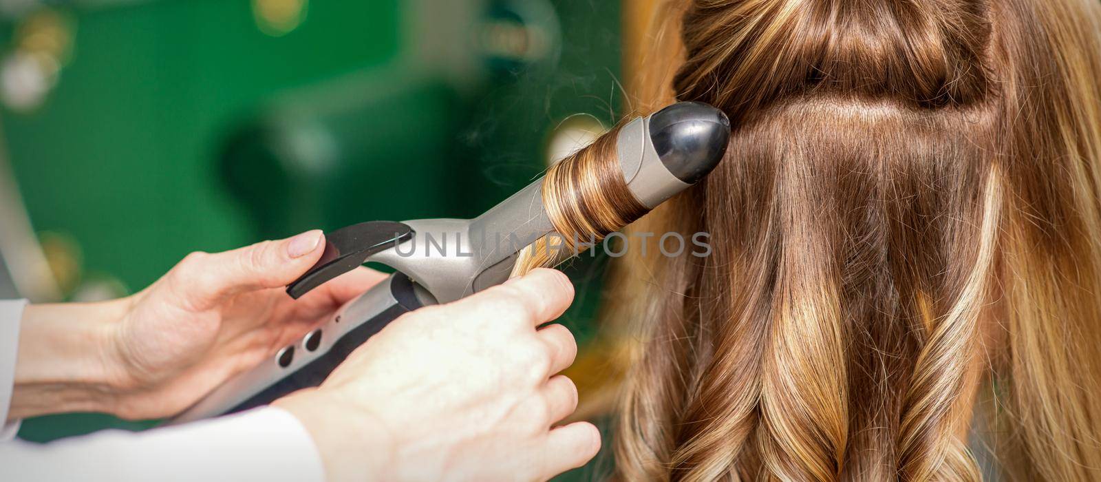 Creating curls with curling irons. Hairdresser makes a hairstyle for a young woman with long red hair in a beauty salon. by okskukuruza