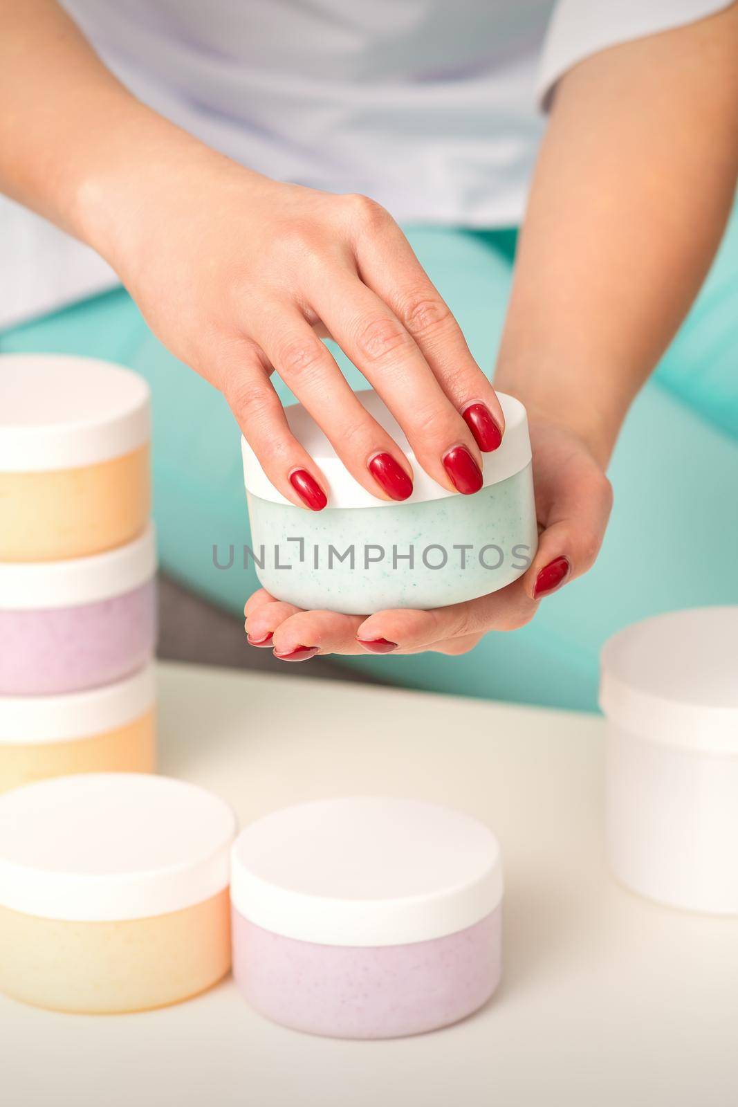 Female hands open a white body cream jar over the table with many jars of cosmetics. by okskukuruza