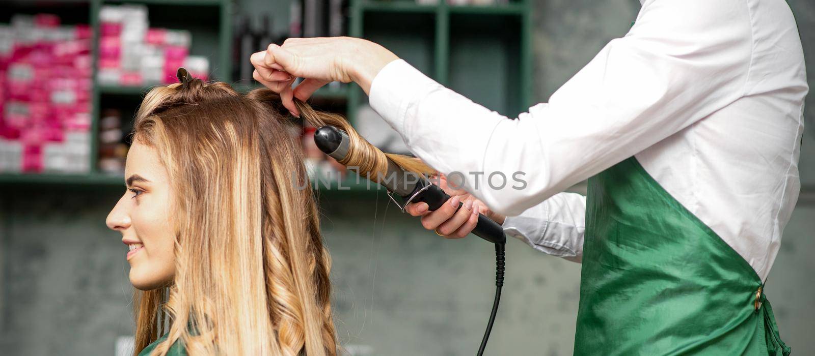 Creating curls with curling irons. Hairdresser makes a hairstyle for a young woman with long red hair in a beauty salon. by okskukuruza