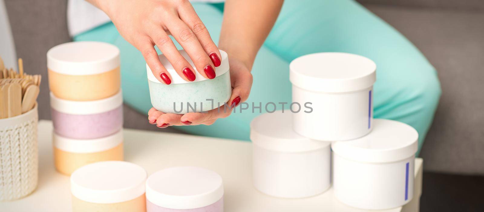 Female hands open a white body cream jar over the table with many jars of cosmetics. by okskukuruza