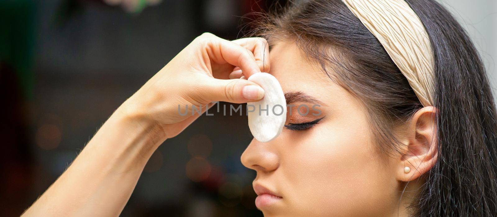 Side view of a makeup artist covers female eye with a cotton pad using airbrush making makeup foundation on her face in a beauty salon