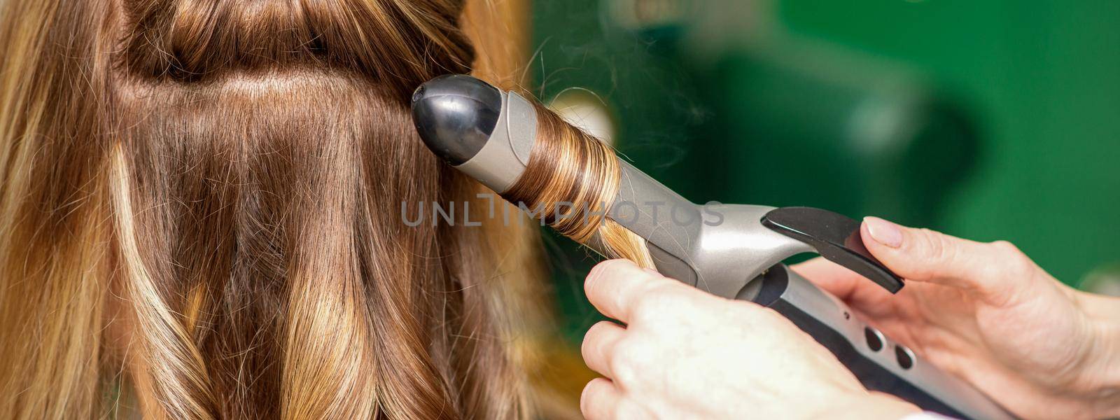 Creating curls with curling irons. Hairdresser makes a hairstyle for a young woman with long red hair in a beauty salon. by okskukuruza