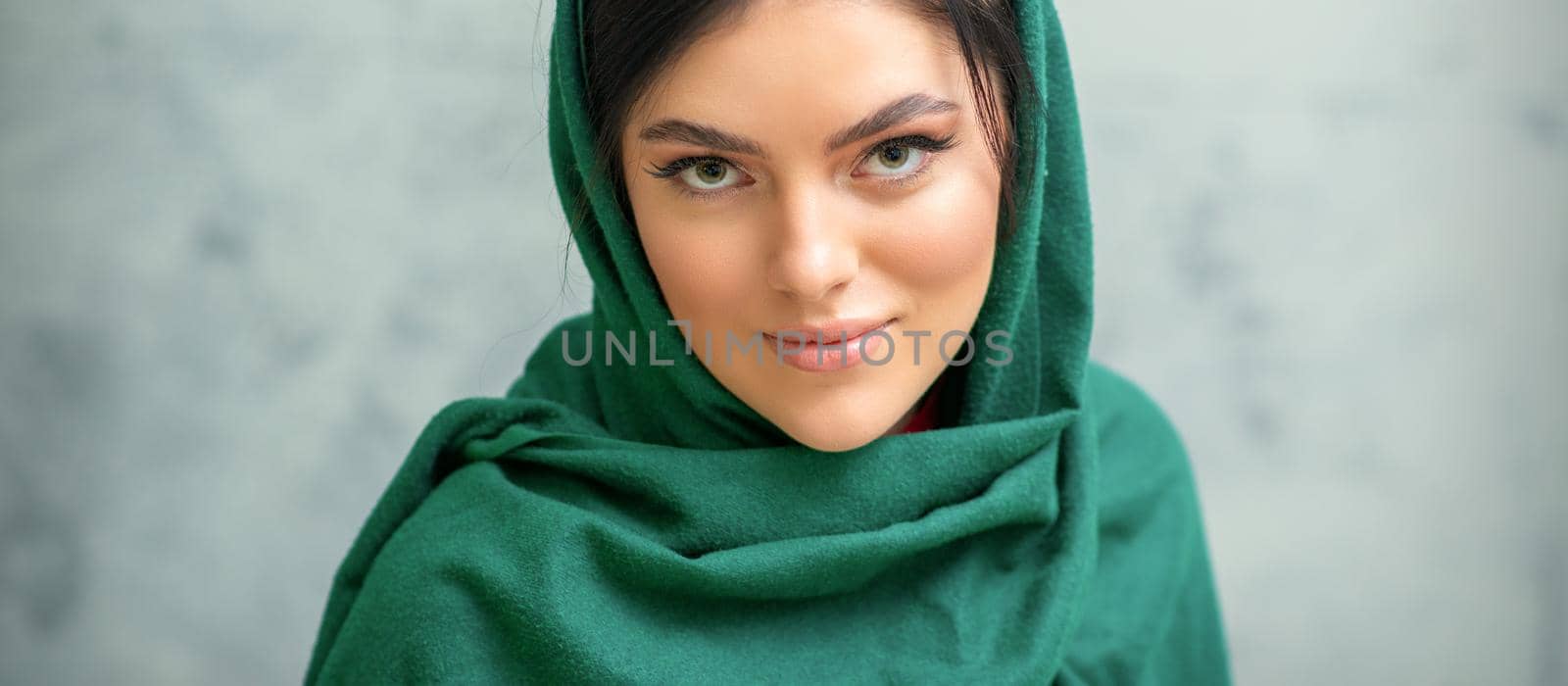 Portrait of a pretty young caucasian woman with makeup in a green headscarf on gray background. by okskukuruza