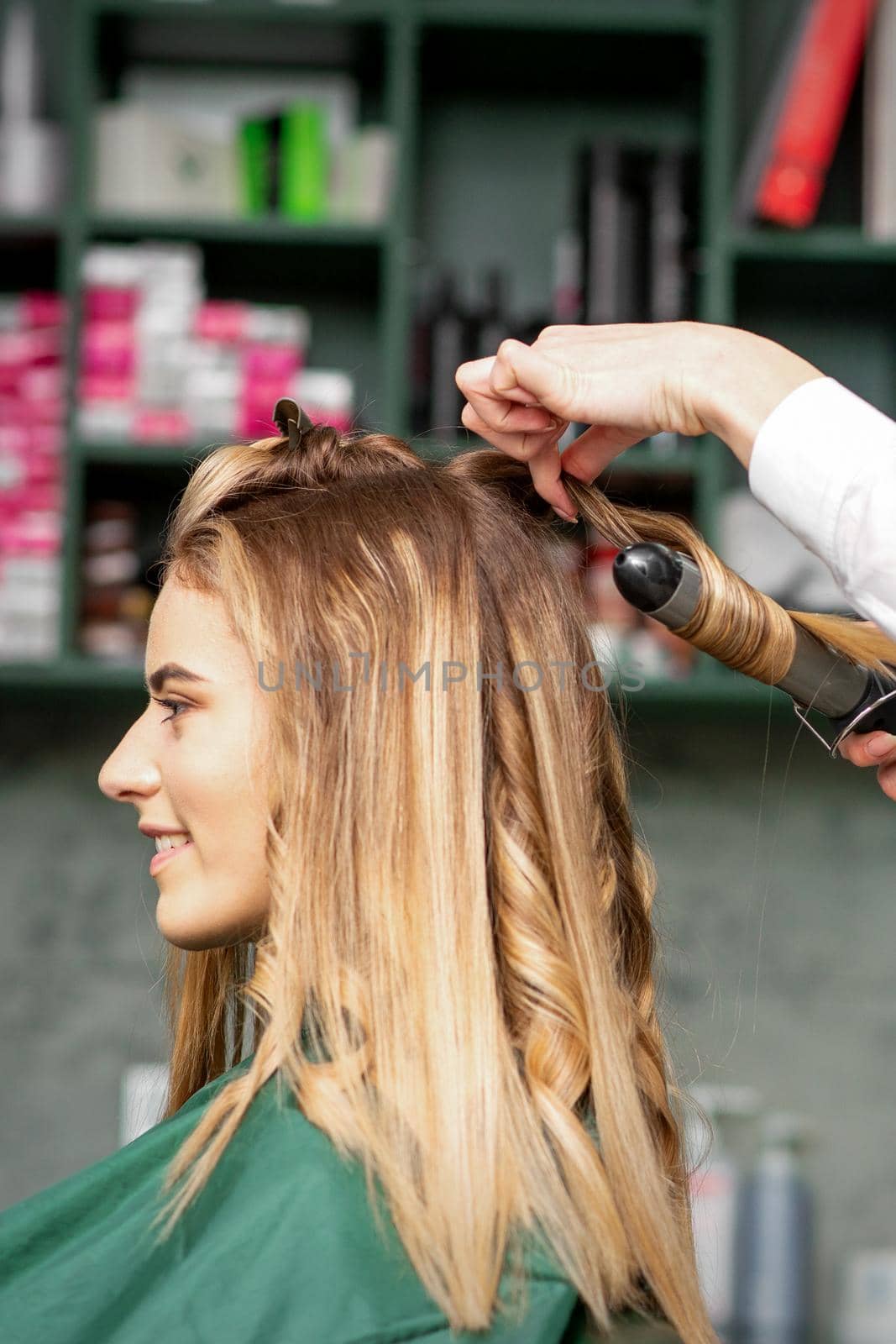 Creating curls with curling irons. Hairdresser makes a hairstyle for a young woman with long red hair in a beauty salon. by okskukuruza