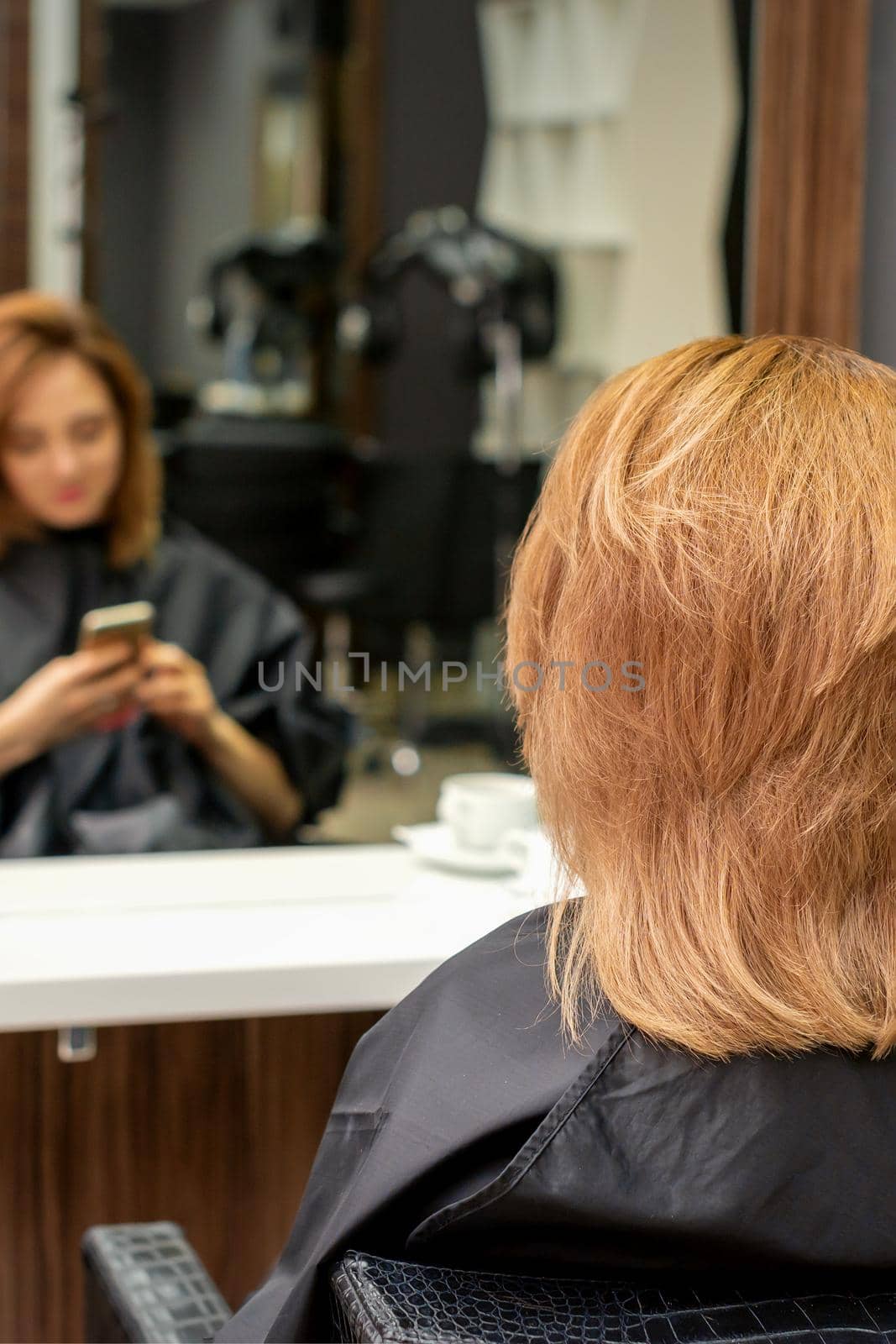 Beautiful young red hair woman using her smartphone and texting sitting in front of a mirror waiting to visit a hairdresser