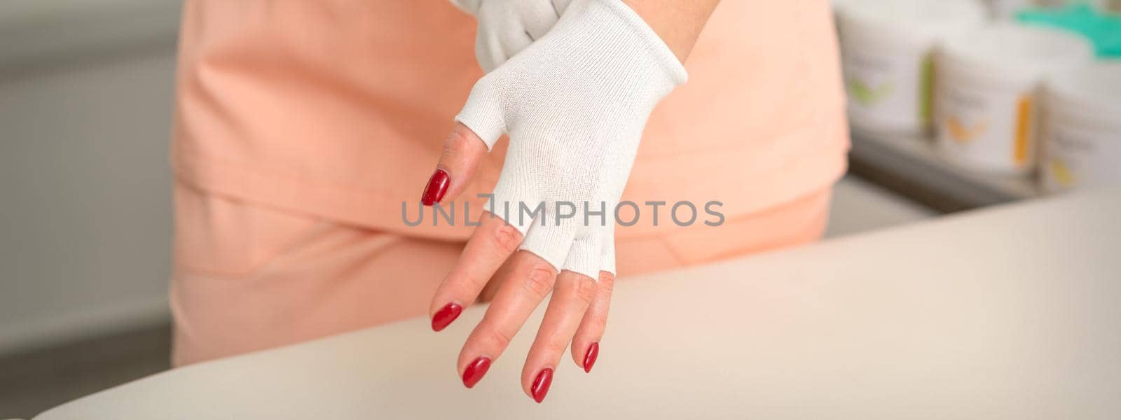 Cosmetician in workwear wearing white bamboo fingerless gloves on her hands in a beauty salon. by okskukuruza