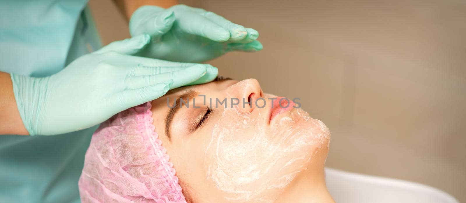 Face peeling at the beautician. Facial treatment. The beautician applies a cleansing face mask to the female patient. by okskukuruza