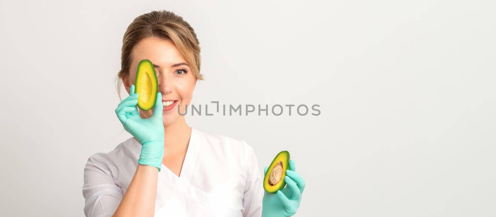 Portrait of young female nutritionist doctor with beautiful smile posing at camera hiding eye behind half avocado on white background, copy space. Benefits of proper nutrition