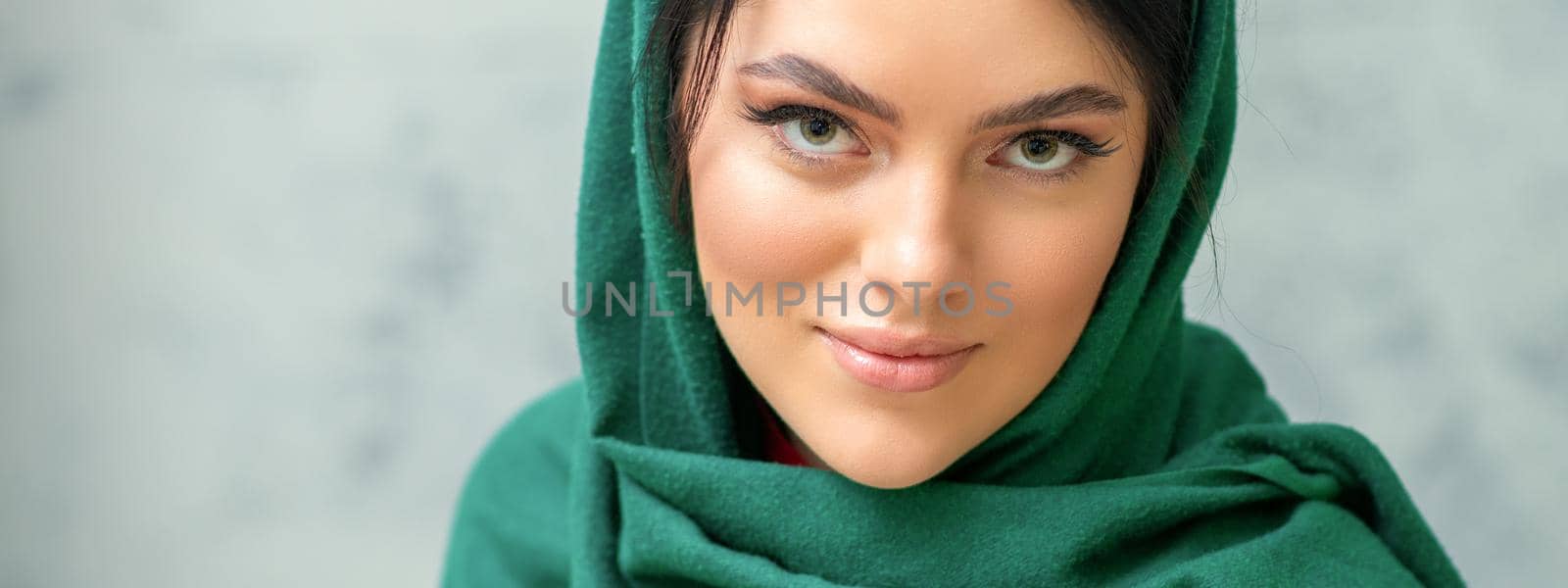 Portrait of a pretty young caucasian woman with makeup in a green headscarf on gray background. by okskukuruza